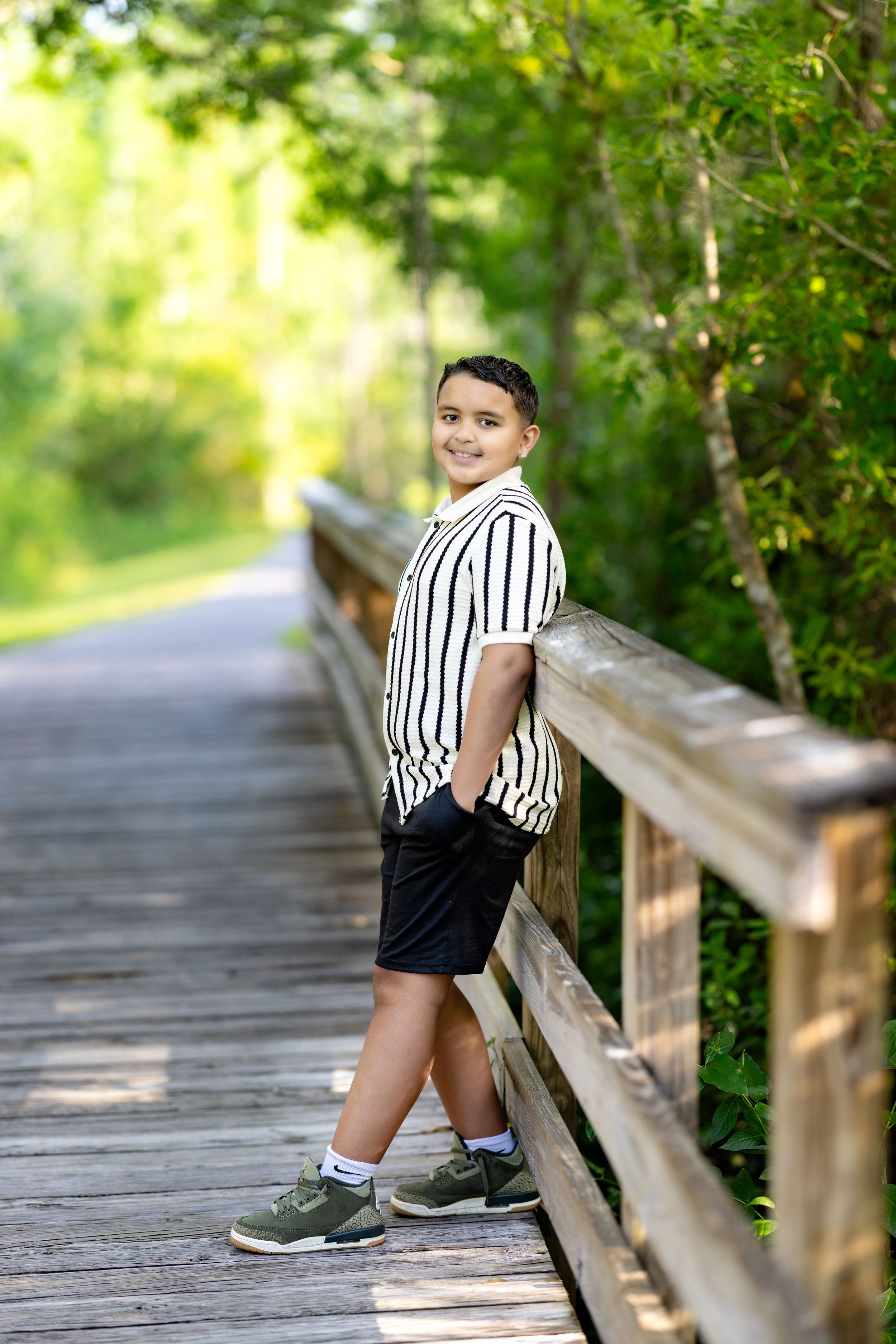 Smiling boy in striped shirt and black shorts leaning on wooden bridge railing surrounded by lush green trees.
