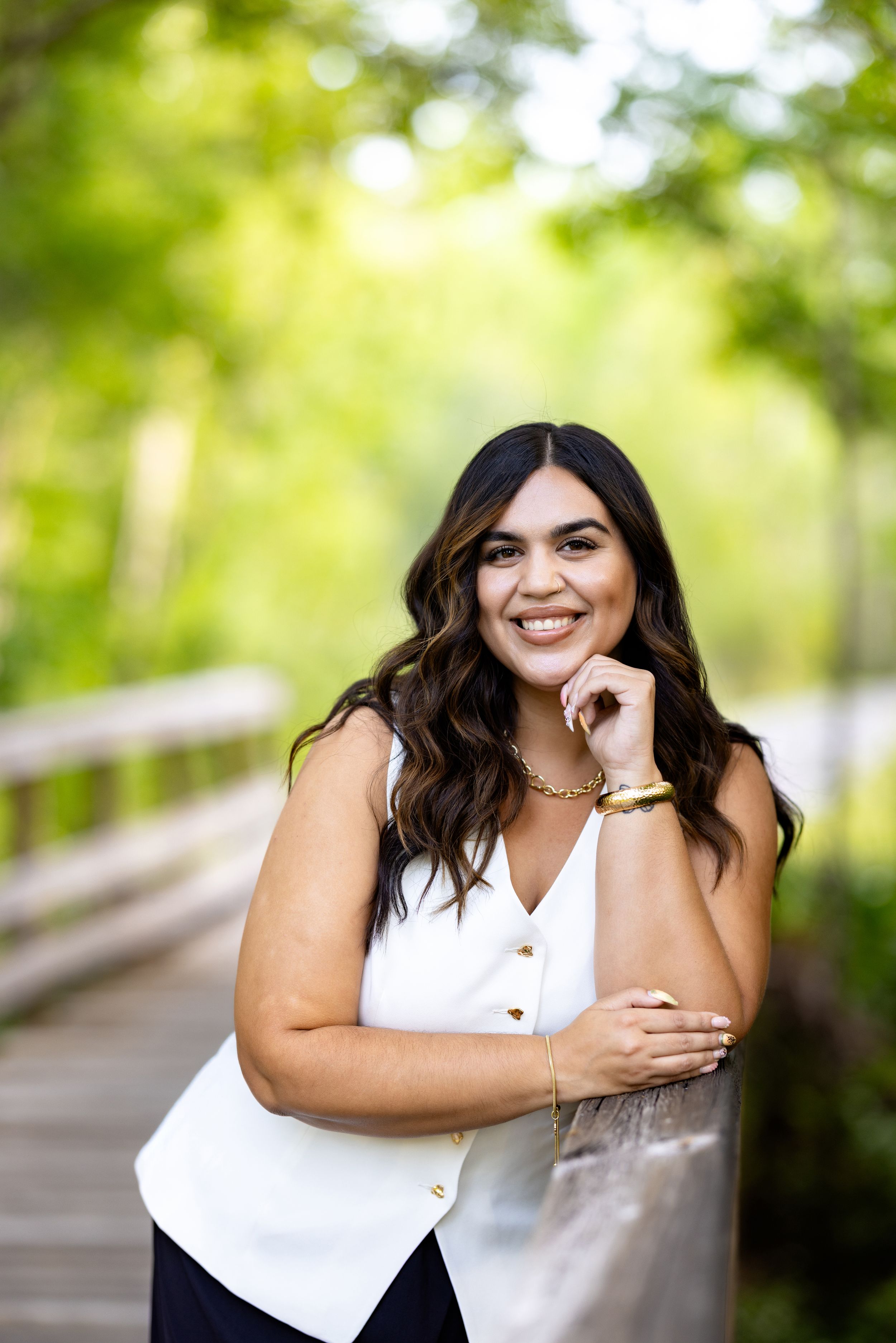 Smiling young woman in white sleeveless top leaning on wooden railing outdoors with lush green trees background.