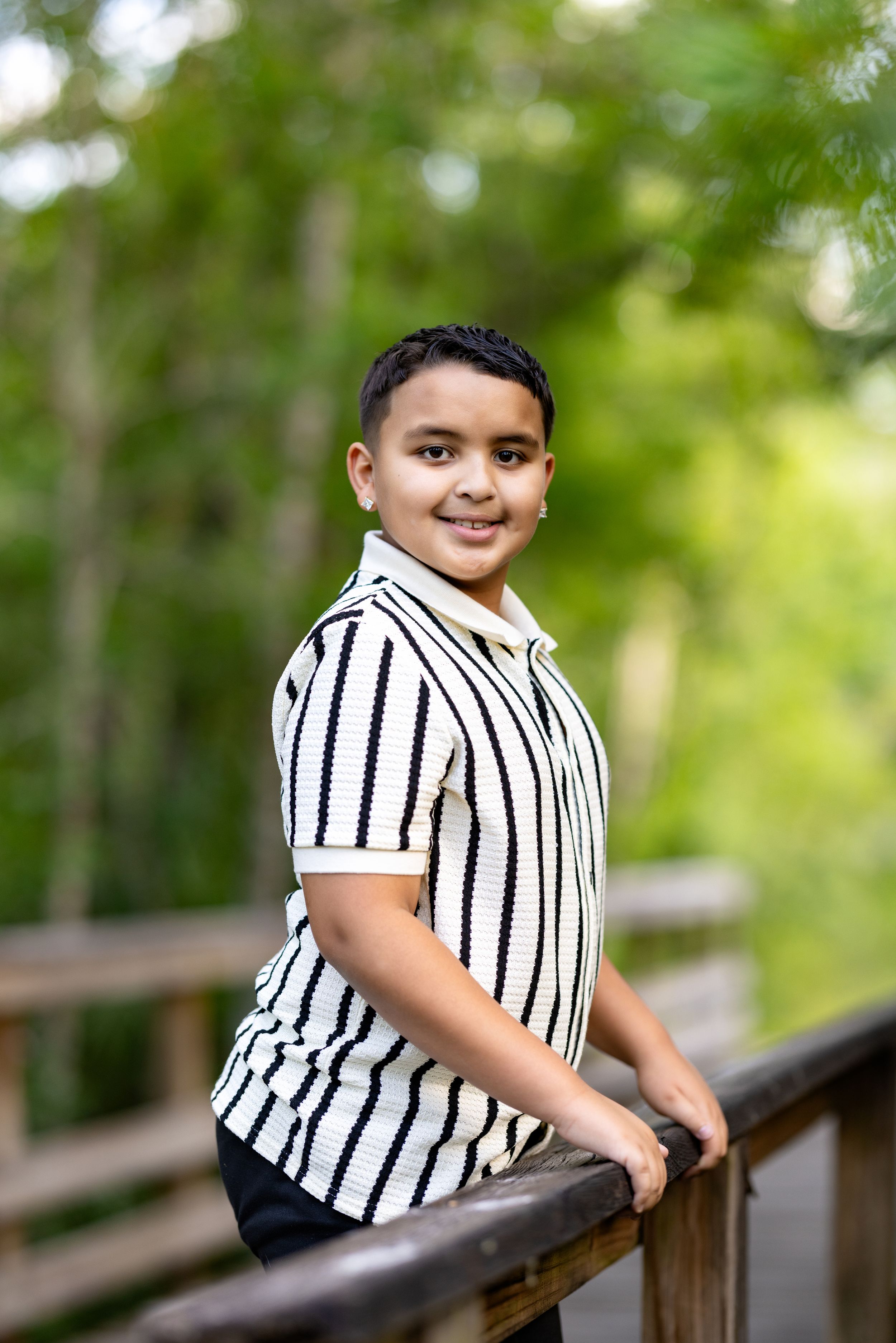 Smiling boy in striped shirt leaning on wooden railing outdoors with lush green trees in background.