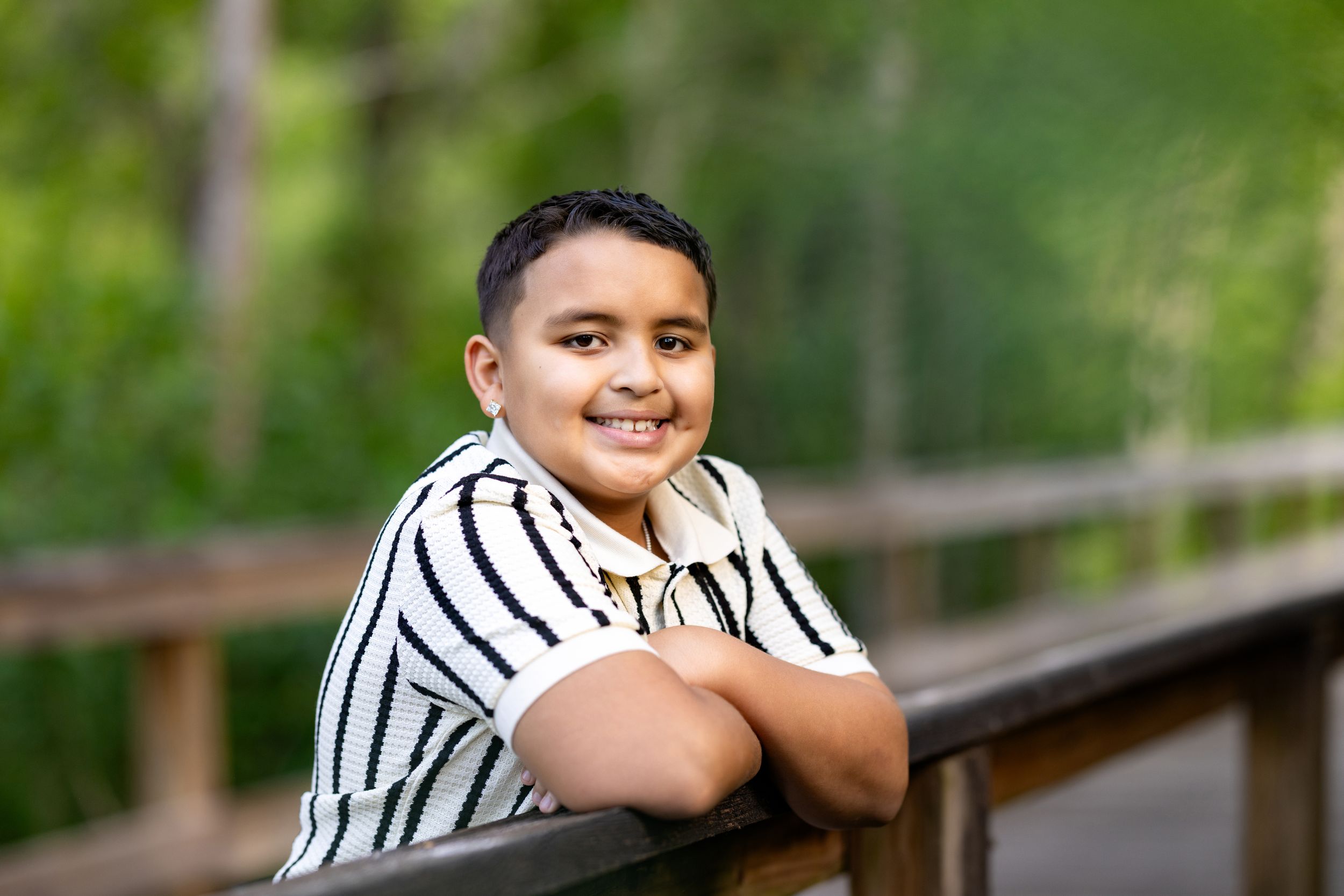 Smiling boy leaning on wooden railing outdoors, wearing striped shirt with green trees in background.