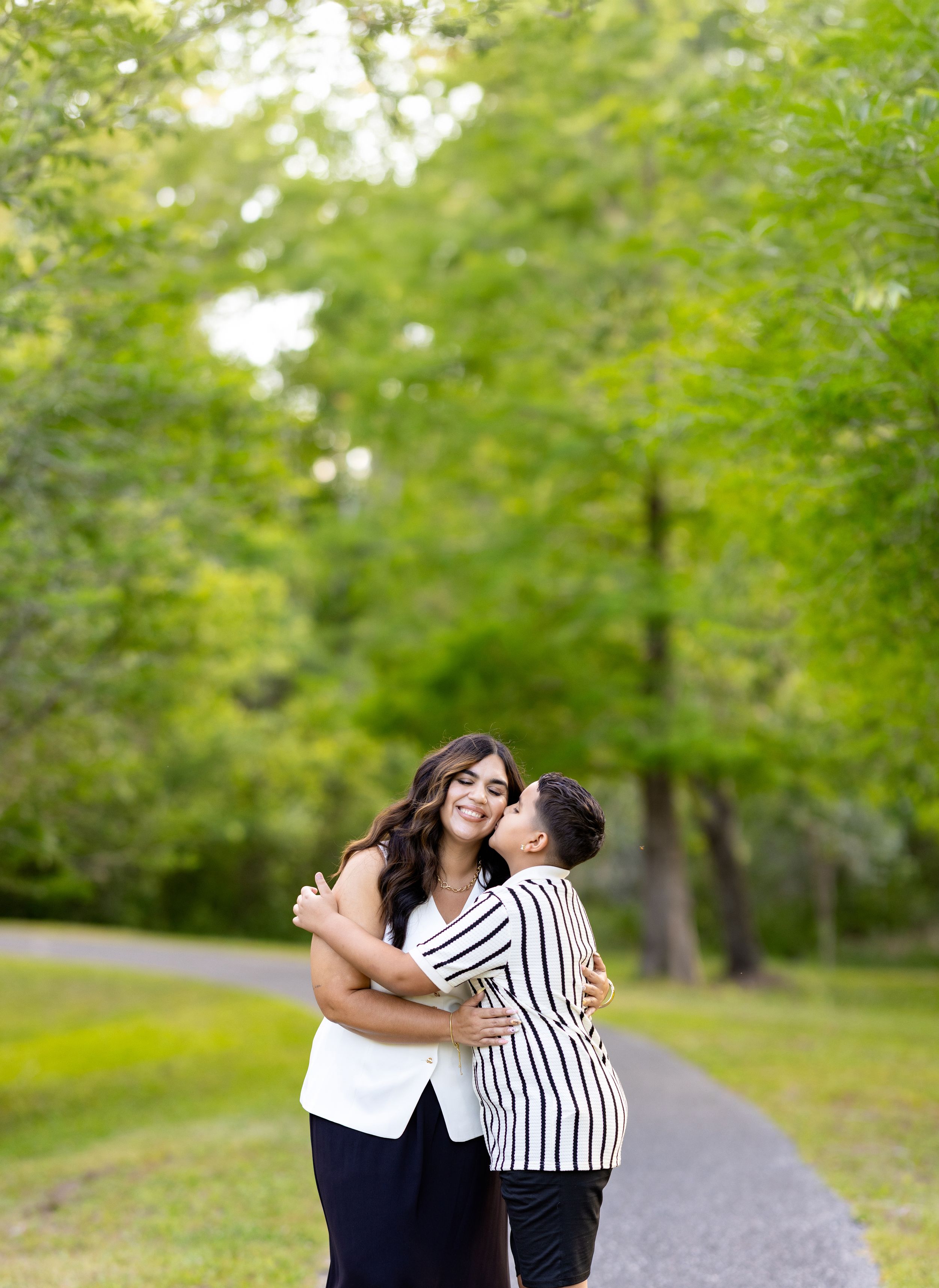 Couple embracing and sharing a kiss on a scenic park path surrounded by lush green trees.