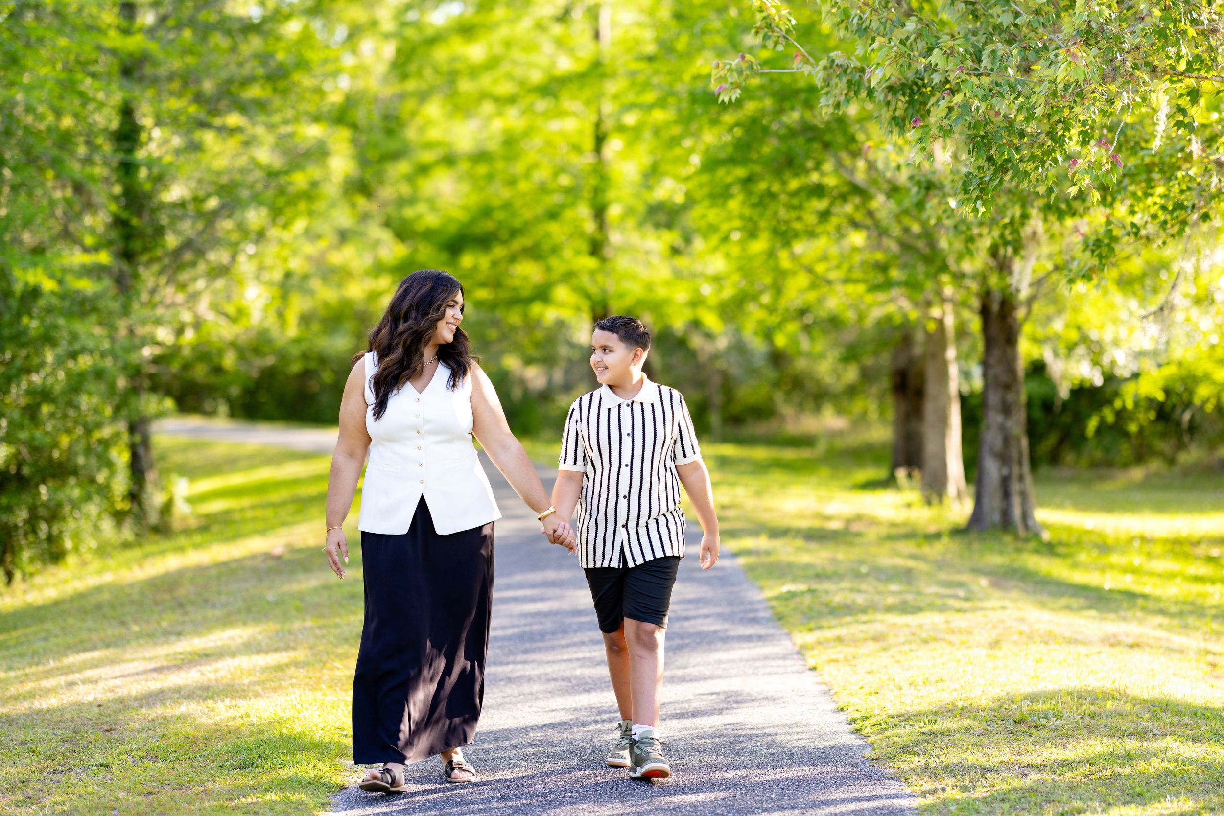 A woman and child walk hand-in-hand on a sunlit park path surrounded by lush green trees.