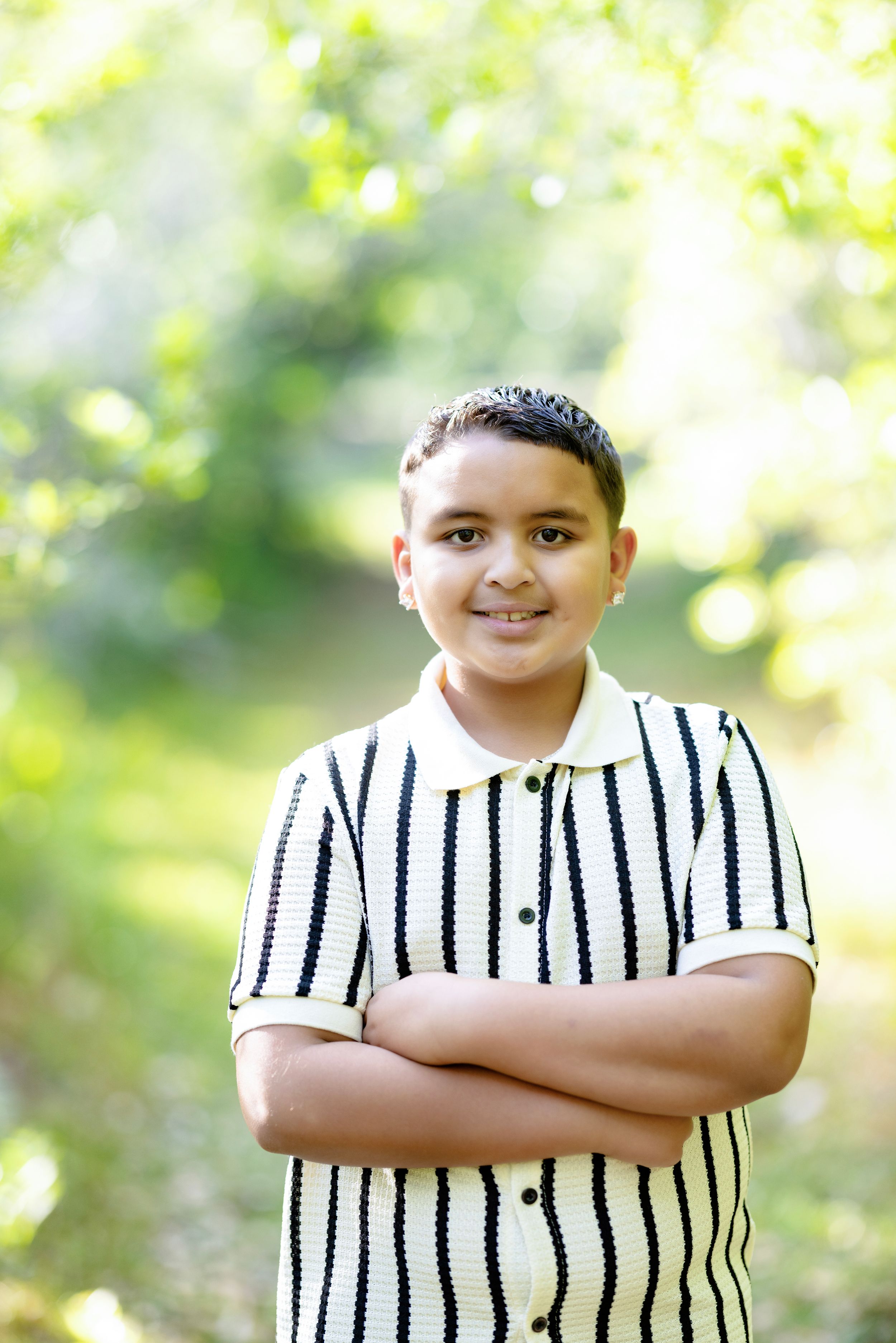 Smiling boy with arms crossed wearing striped shirt outdoors in green nature setting.