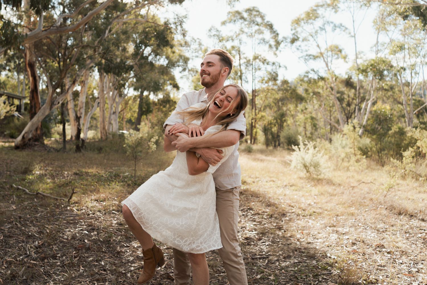 Happy couple embracing and laughing outdoors in sunlit eucalyptus forest, woman in white dress lifted by man couples photoshoot in rouse hil
