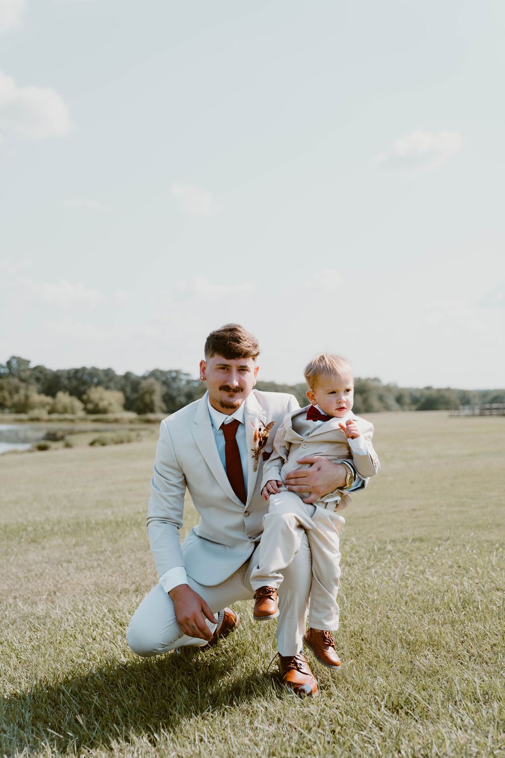 Groom in light gray suit holds young boy in matching outfit on green lawn at outdoor wedding venue.