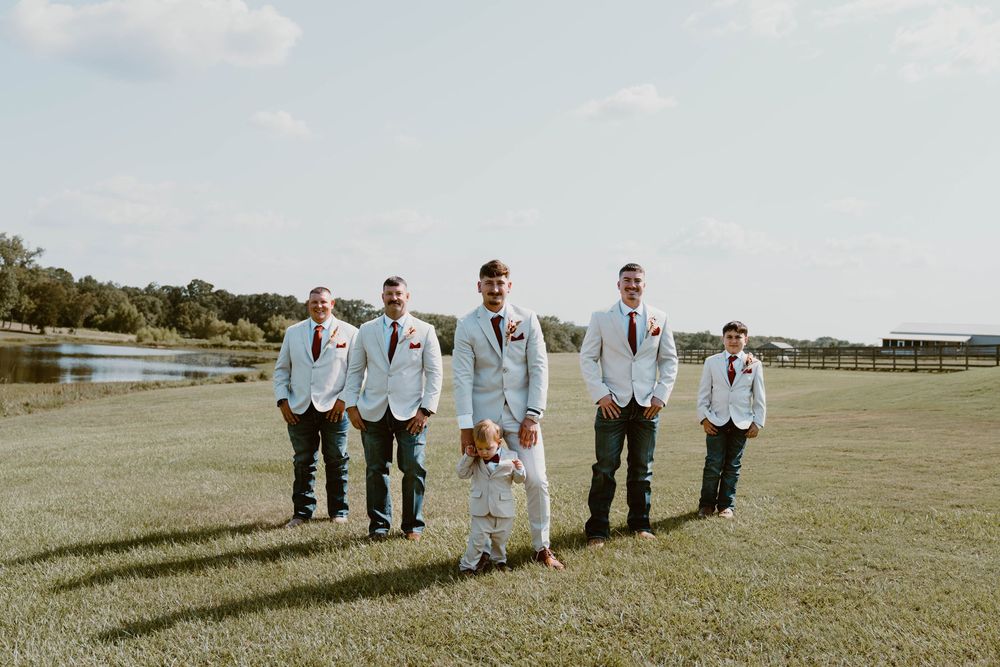 Groomsmen in light blue suits with dark ties stand on green lawn with a dog at an outdoor wedding venue.