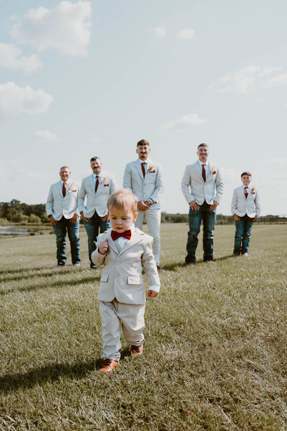 Cute toddler ring bearer in white suit leads groomsmen across a sunny green field at an outdoor wedding.