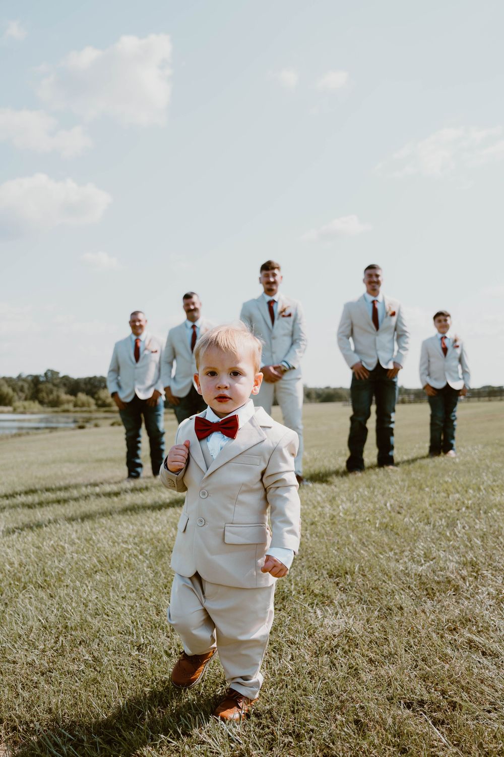 Adorable toddler in a beige suit and red bow tie leads groomsmen across a green lawn at an outdoor wedding.