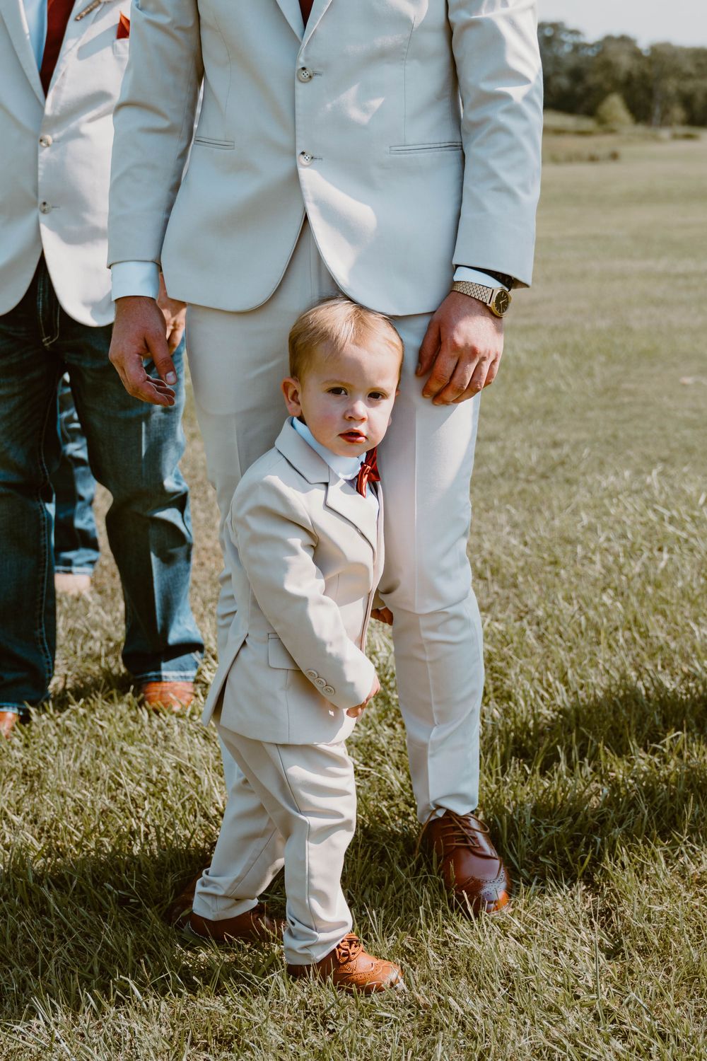 Adorable toddler ring bearer in a white suit stands beside a groomsman in matching attire on a green lawn.