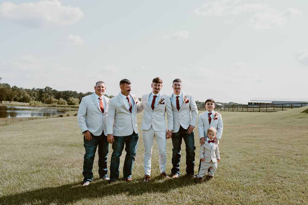 Groomsmen in light blue suits with red ties stand on green lawn with a young ring bearer at an outdoor wedding.