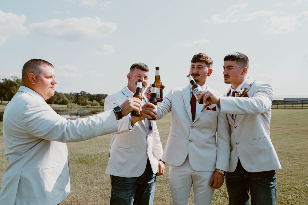 Four groomsmen in light suits toasting with beer bottles outdoors on a sunny wedding day.