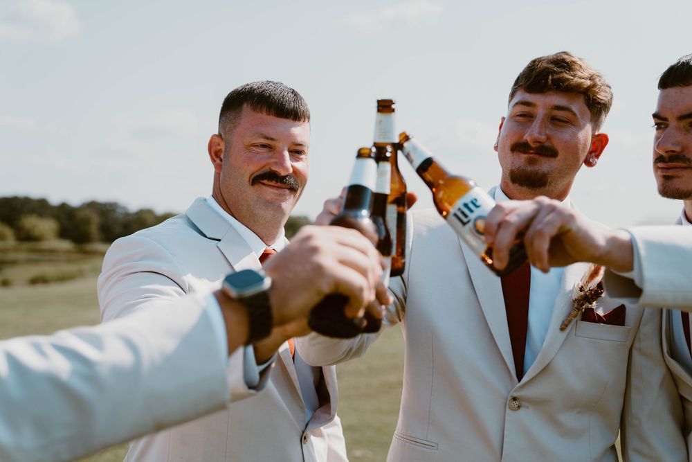 Groomsmen in white suits toasting with beer bottles outdoors at a wedding celebration.