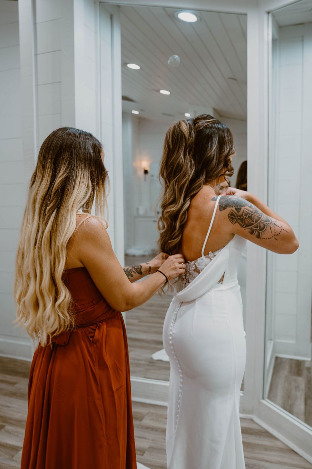 Bridesmaid in rust dress fastening buttons on brides white wedding gown before ceremony.