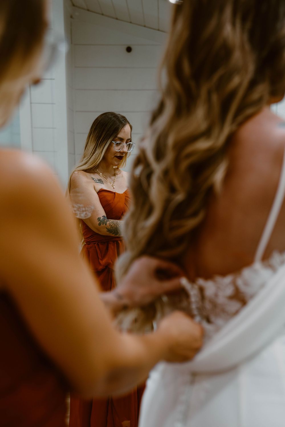 Mirror reflection of tattooed bridesmaid in orange dress helping bride with wedding gown buttons.