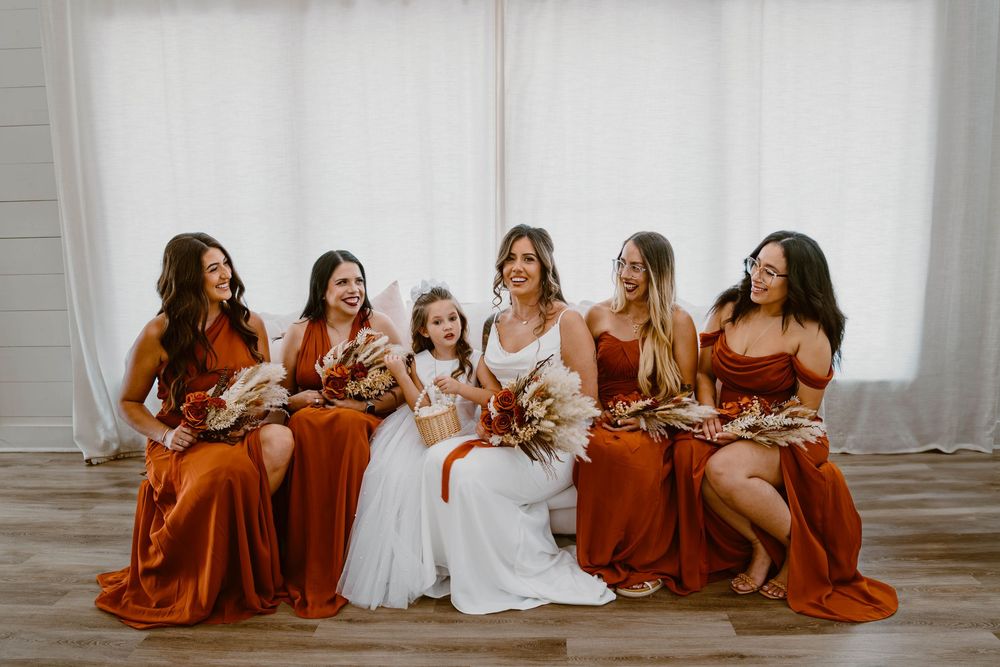 Bride in white gown with bridesmaids in rust orange dresses and flower girl seated together holding bouquets.