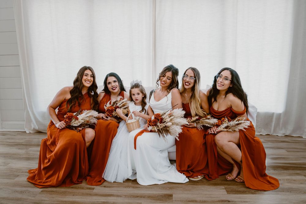 Bride in white gown seated with four bridesmaids in rust orange dresses and flower girl laughing.