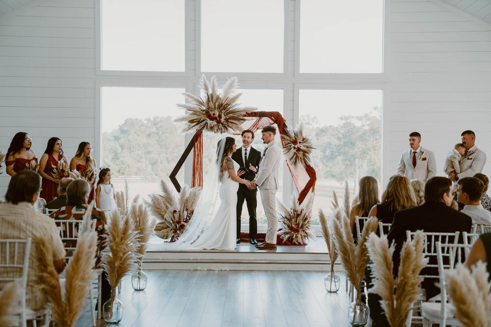 Elegant indoor barn wedding ceremony with boho decor, pampas grass, and a geometric floral arch backdrop.