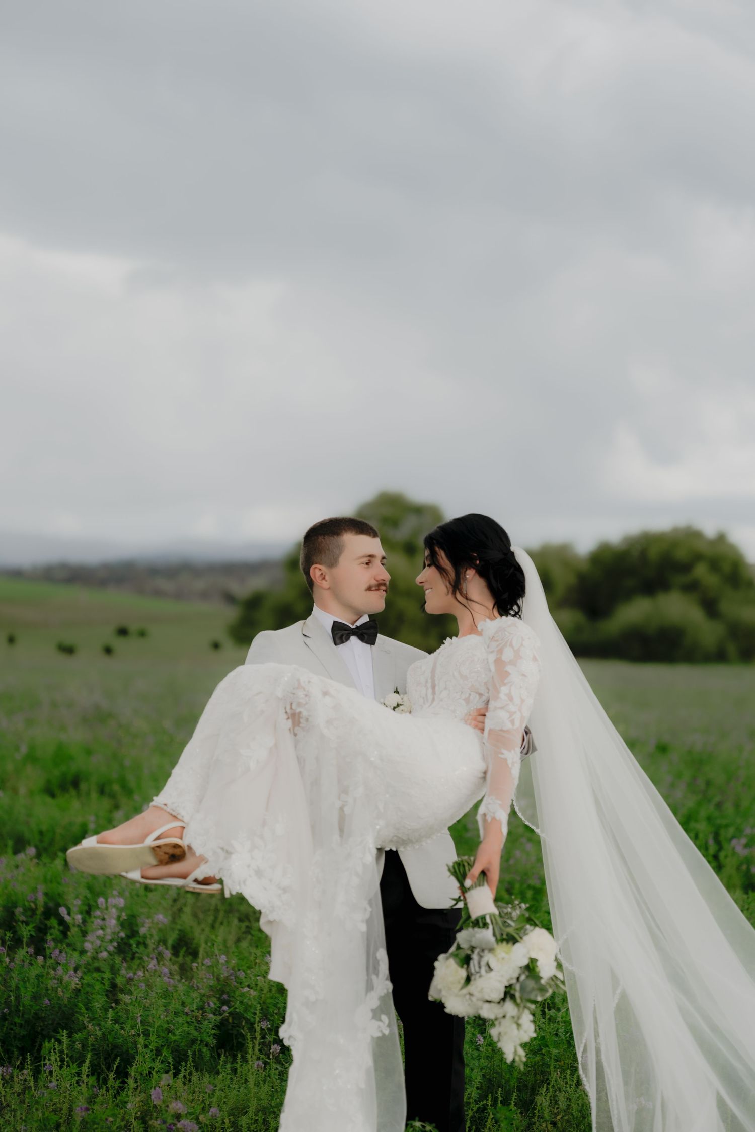 Groom in black tux carries bride in white lace gown through green lavender field on cloudy wedding day.