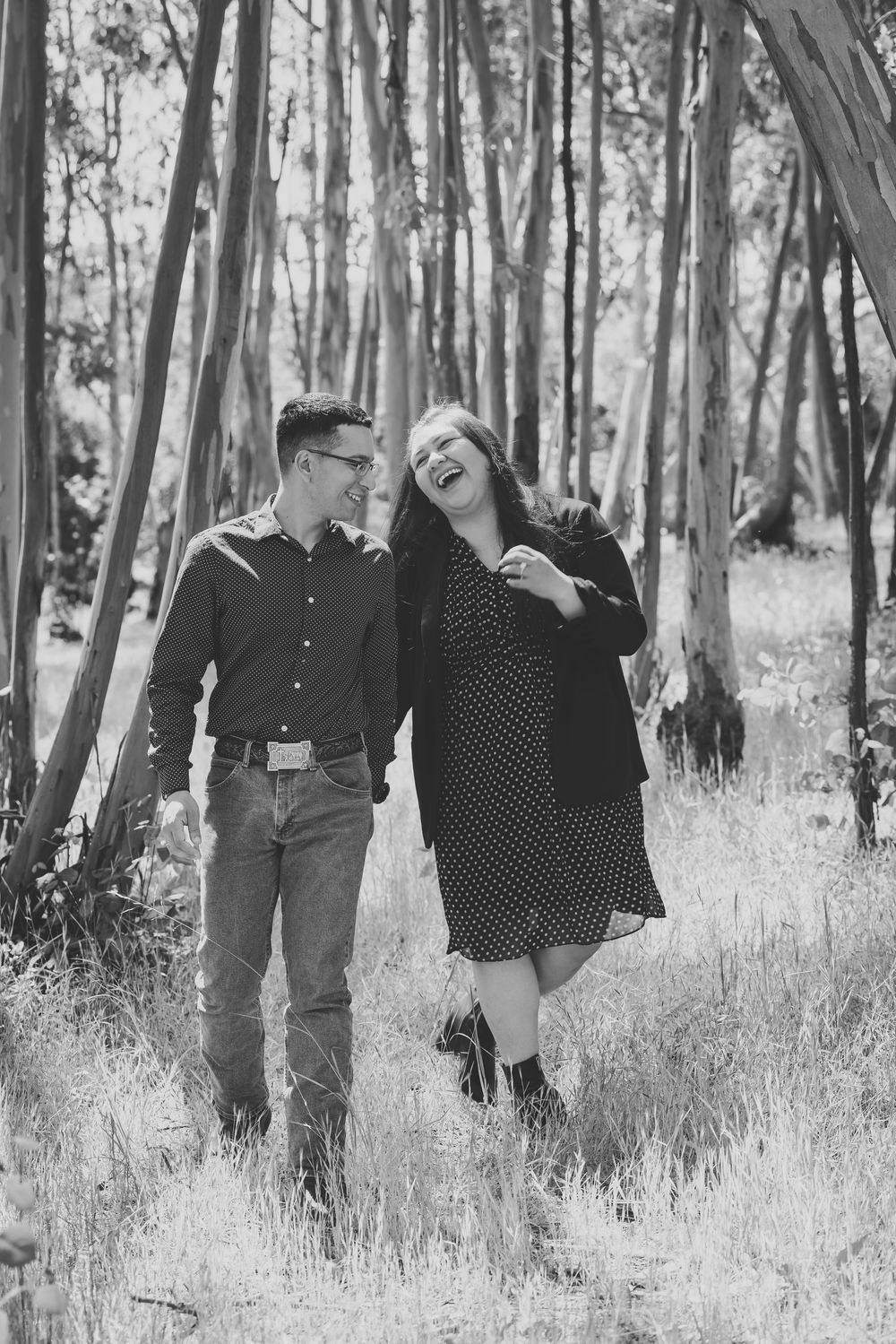 A laughing couple walks together through a forest in this black and white engagement photo.