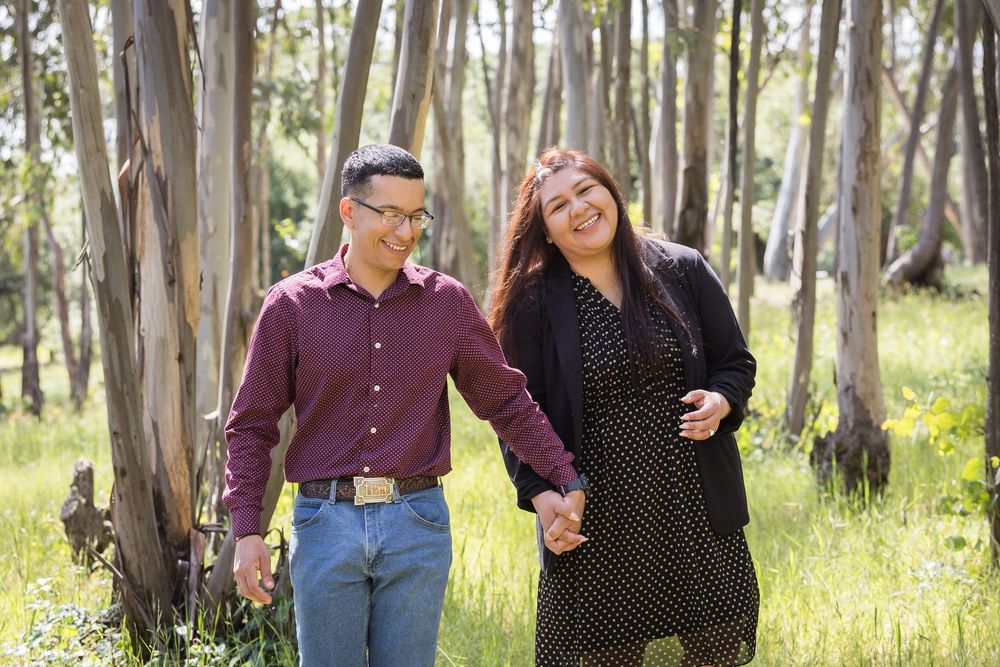 Couple holding hands and smiling while walking through a lush green forest with tall eucalyptus trees.