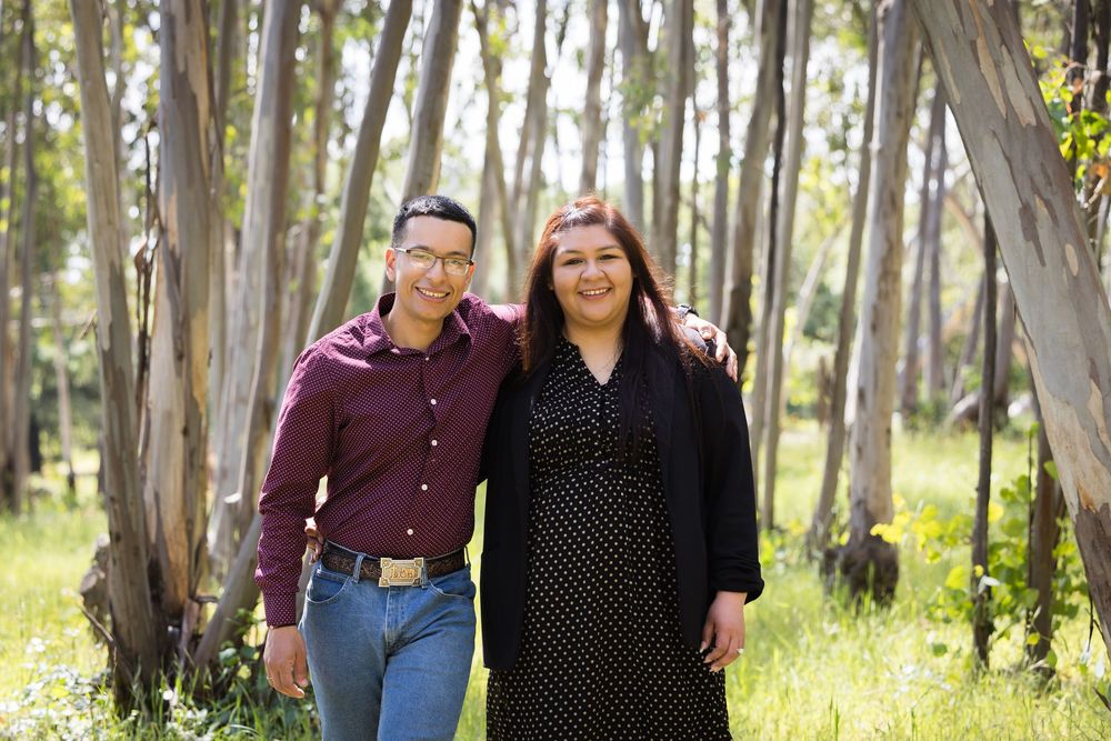 A smiling couple poses together in a sunlit eucalyptus forest, man in maroon shirt and woman in polka dot dress.