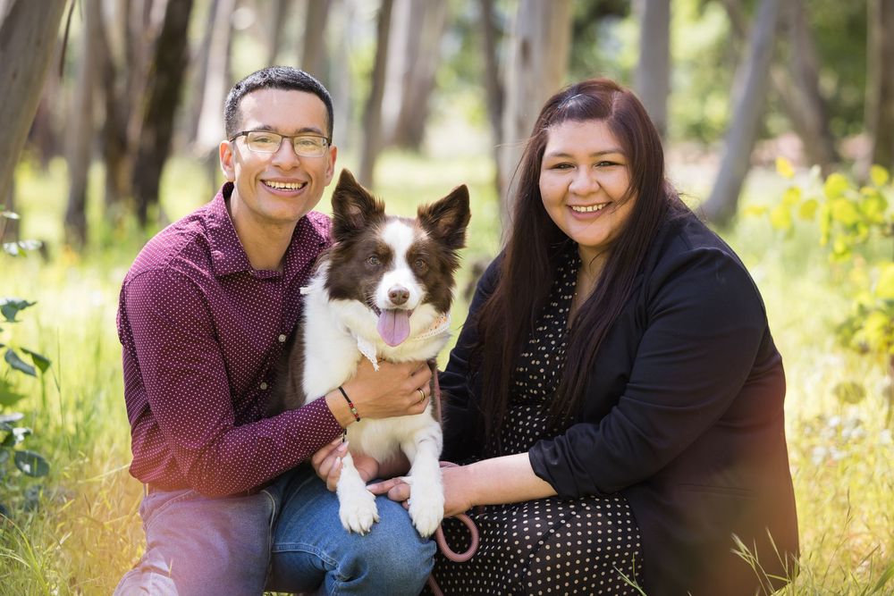 A smiling couple kneeling outdoors in a sunlit forest, holding a black and white Border Collie dog.