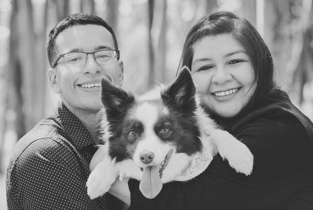 Black and white photo of smiling couple holding a Border Collie dog outdoors among trees.