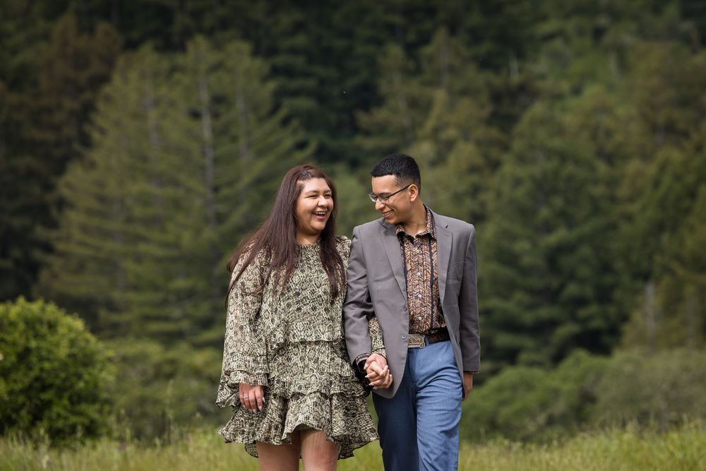 A couple walks hand-in-hand through a lush green meadow, smiling at each other with tall evergreen trees behind them.