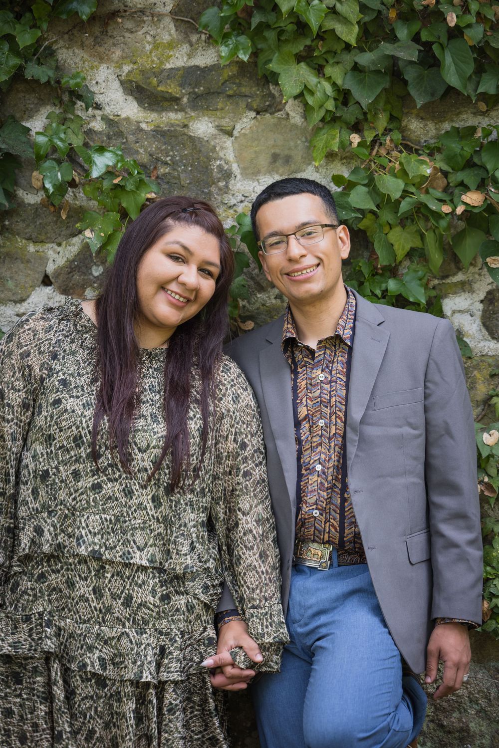 Smiling couple posing together against a stone wall with ivy, woman in floral dress, man in gray blazer.