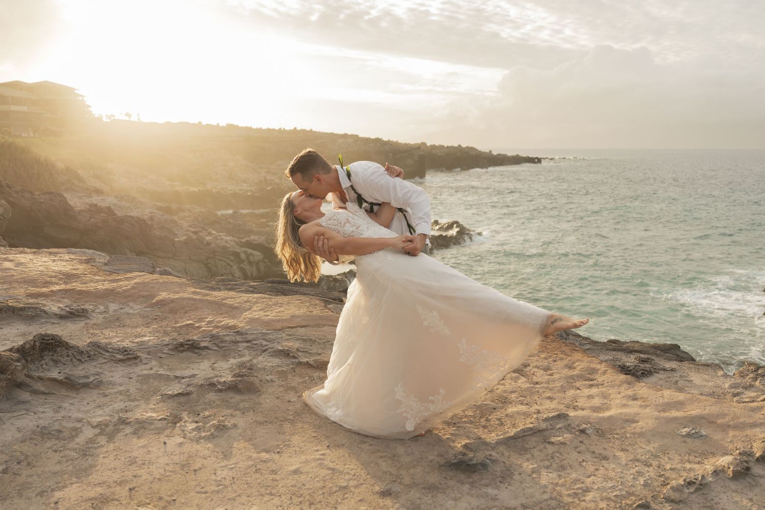 Couple sharing a romantic dip kiss in wedding attire on rocky cliffs at sunset by the ocean.