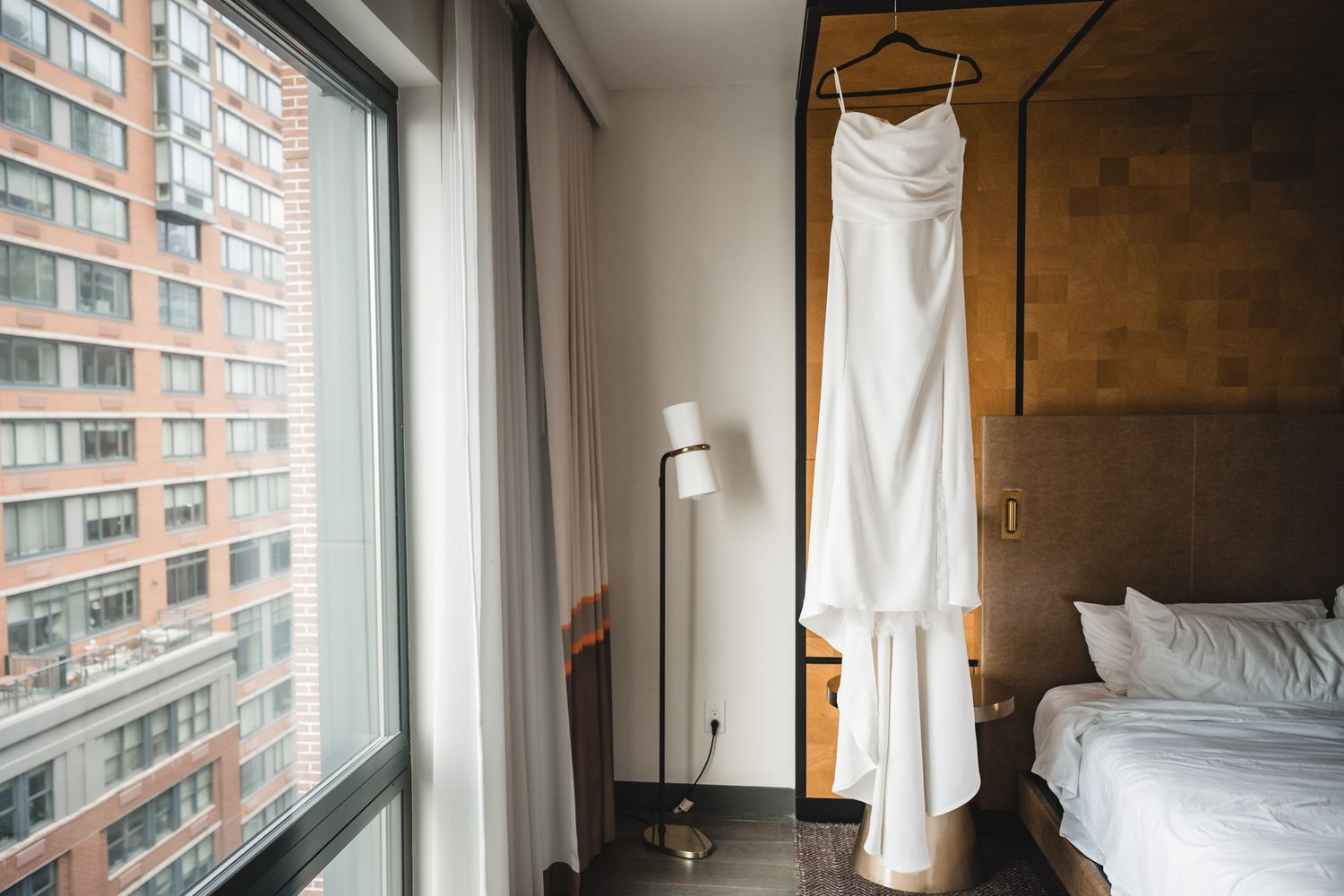Elegant white wedding dress hanging in a modern hotel room with floor-to-ceiling windows and city views.