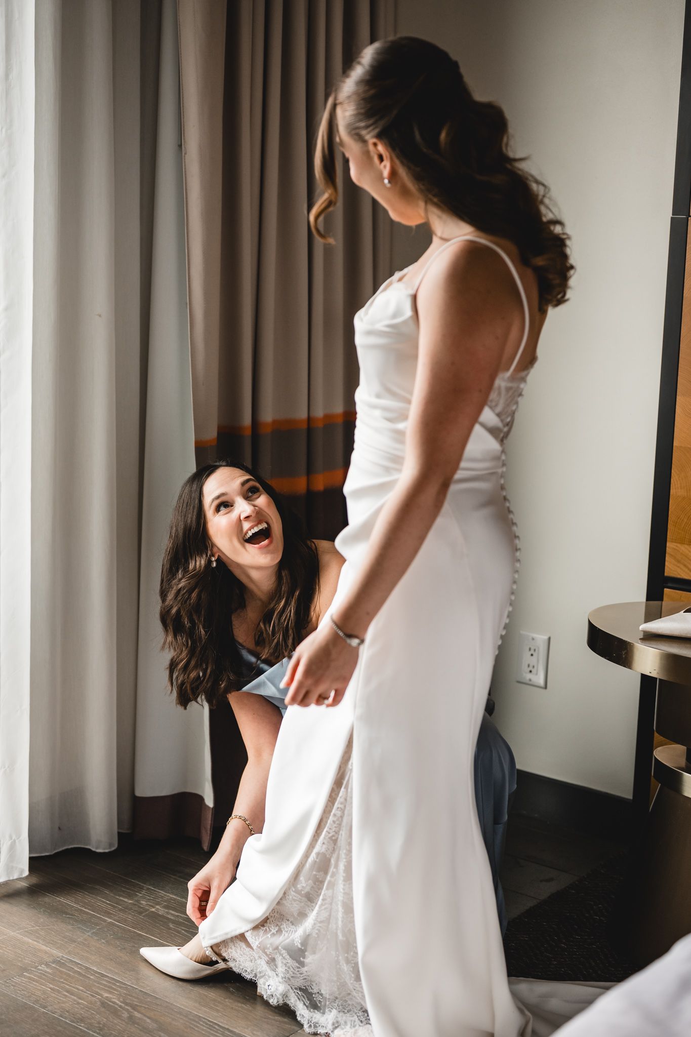 Bridesmaid kneeling to help bride put on white heels while laughing in a hotel room on wedding day.