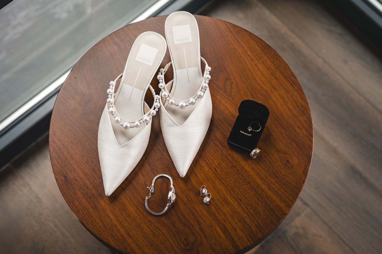 White pearl-embellished bridal heels displayed on wooden table with ring box, earrings, and bracelet.