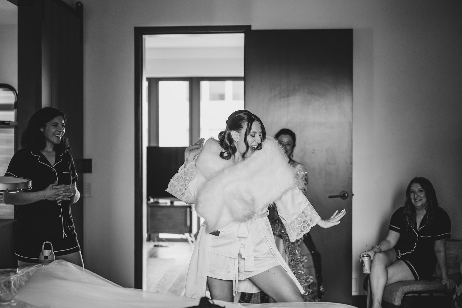 Bridesmaids in robes hugging and laughing together in a hotel room during bridal preparations in black and white.