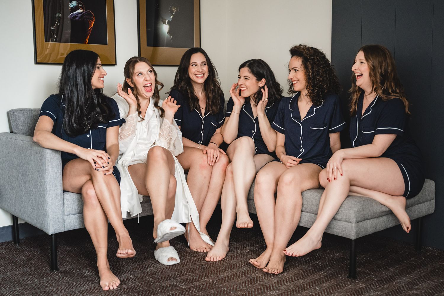 Six women in matching navy pajamas laugh together on a gray couch during a bridal party getting-ready session.