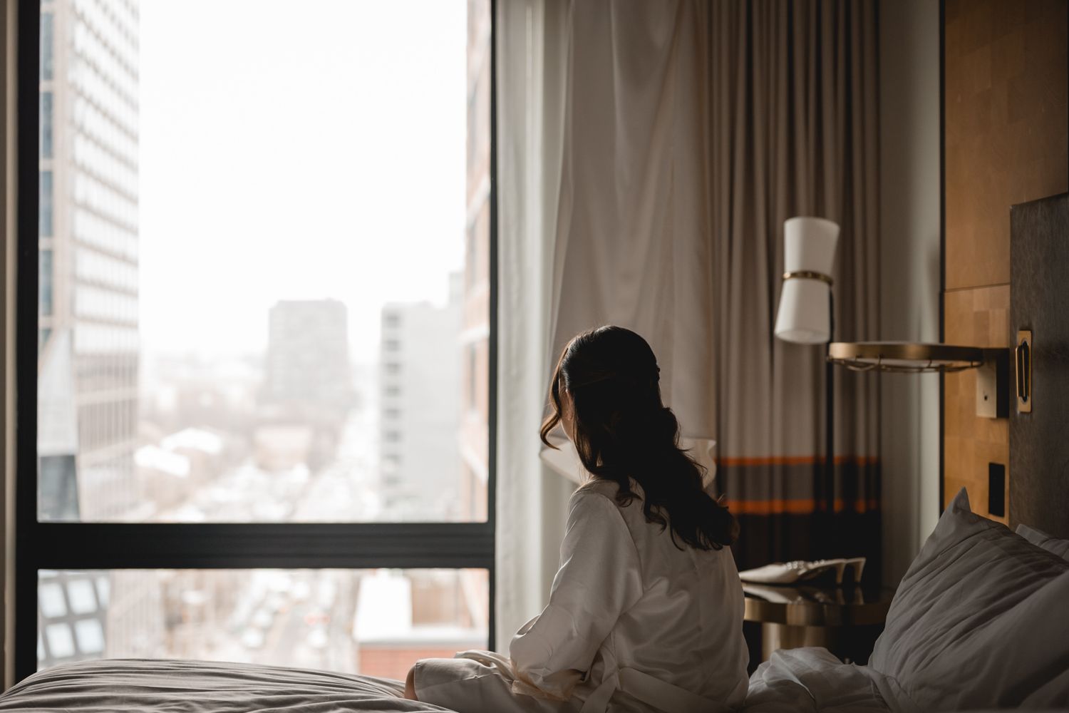Woman in white robe sitting on hotel bed gazing at city skyline through large window with sheer curtains.