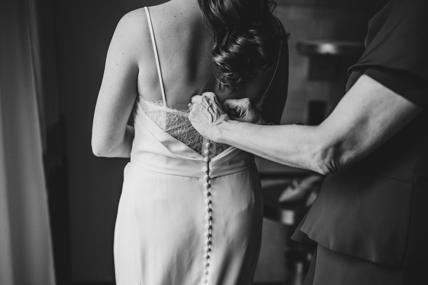 Black and white photo of a bride having her wedding dress buttoned up the back by a helper.