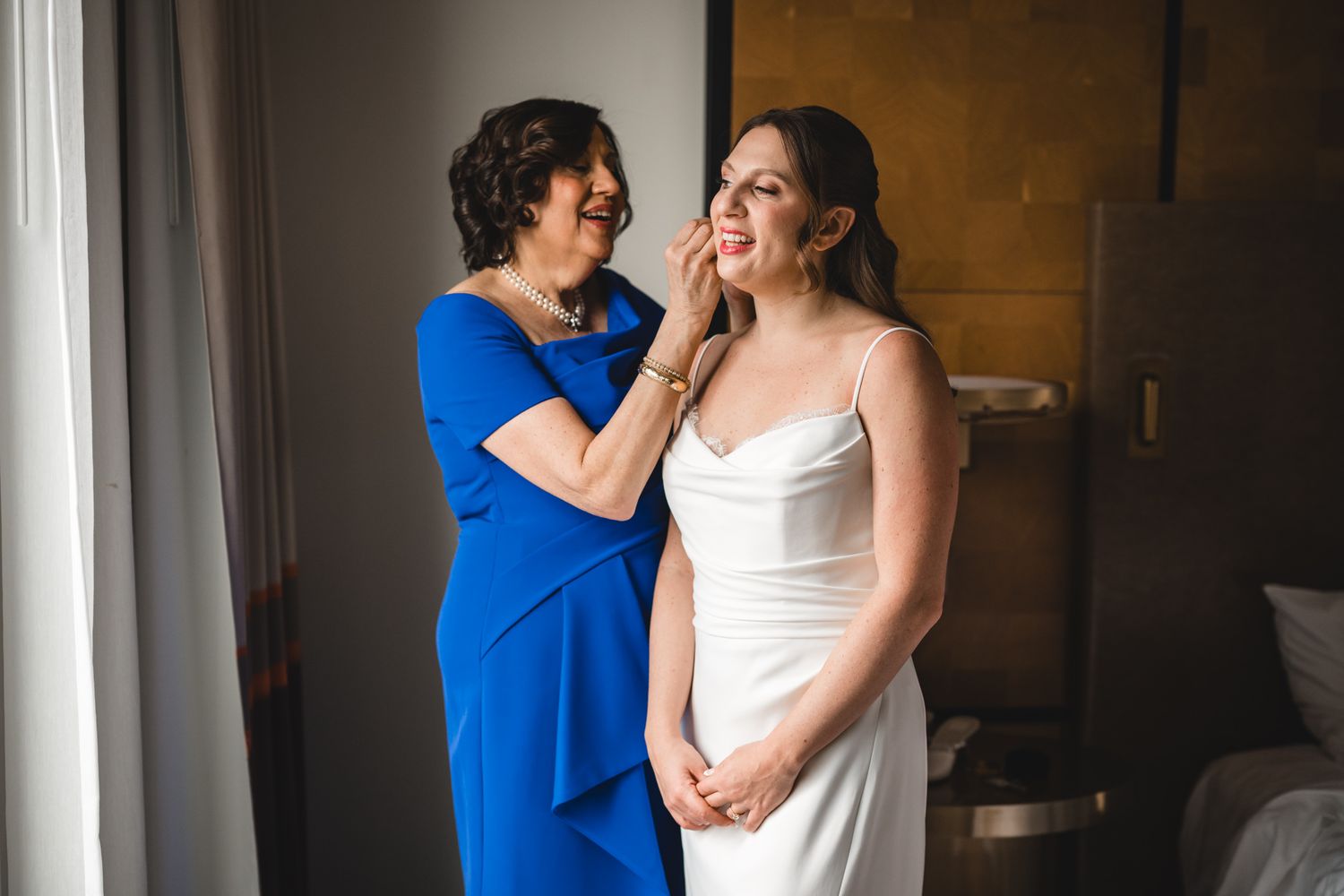 Bride in white gown gets makeup touched up by woman in blue dress during wedding preparations.