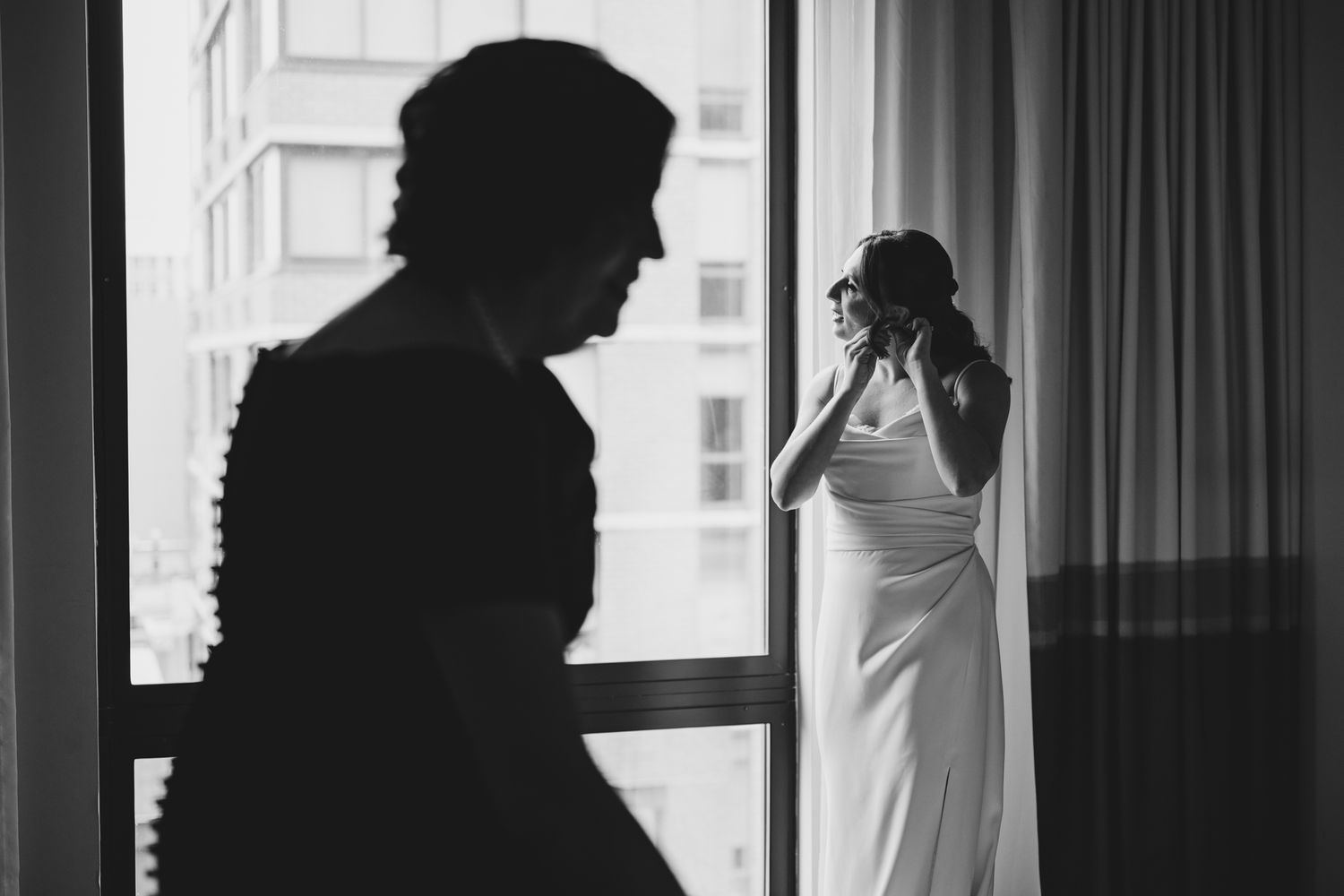 Black and white photo of a bride in a white gown adjusting her hair near a window, with a silhouette in the foreground.