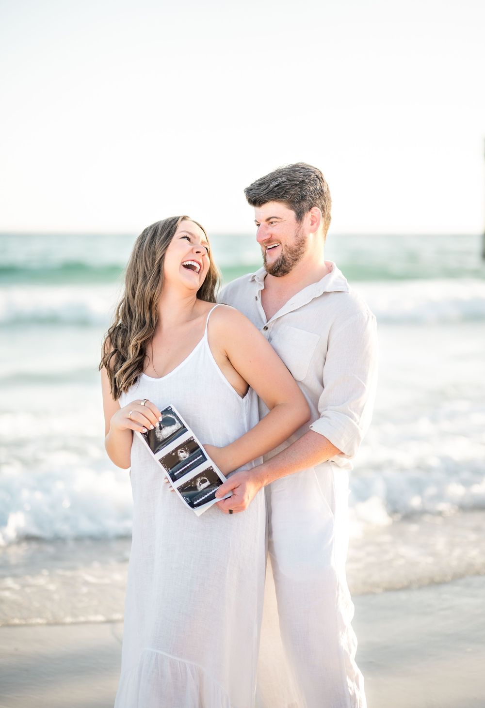 Happy couple in white outfits holding ultrasound photos on a sunny beach, sharing a joyful pregnancy announcement moment.