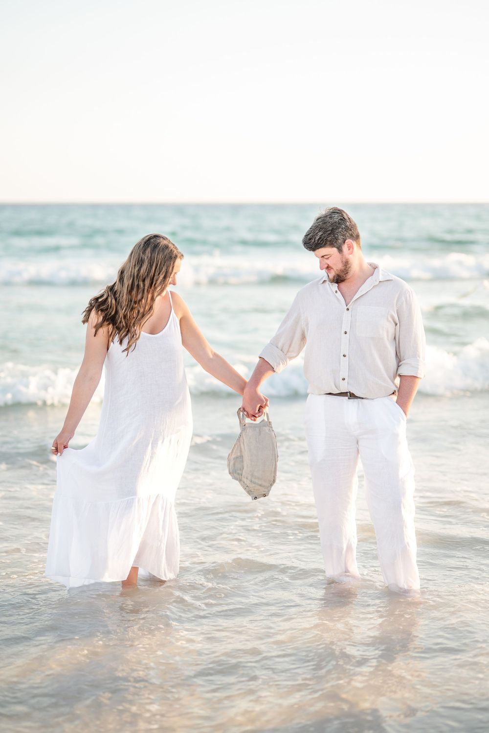 Couple holding hands wading in ocean waves at beach, dressed in white and beige outfits, carrying a woven bag.