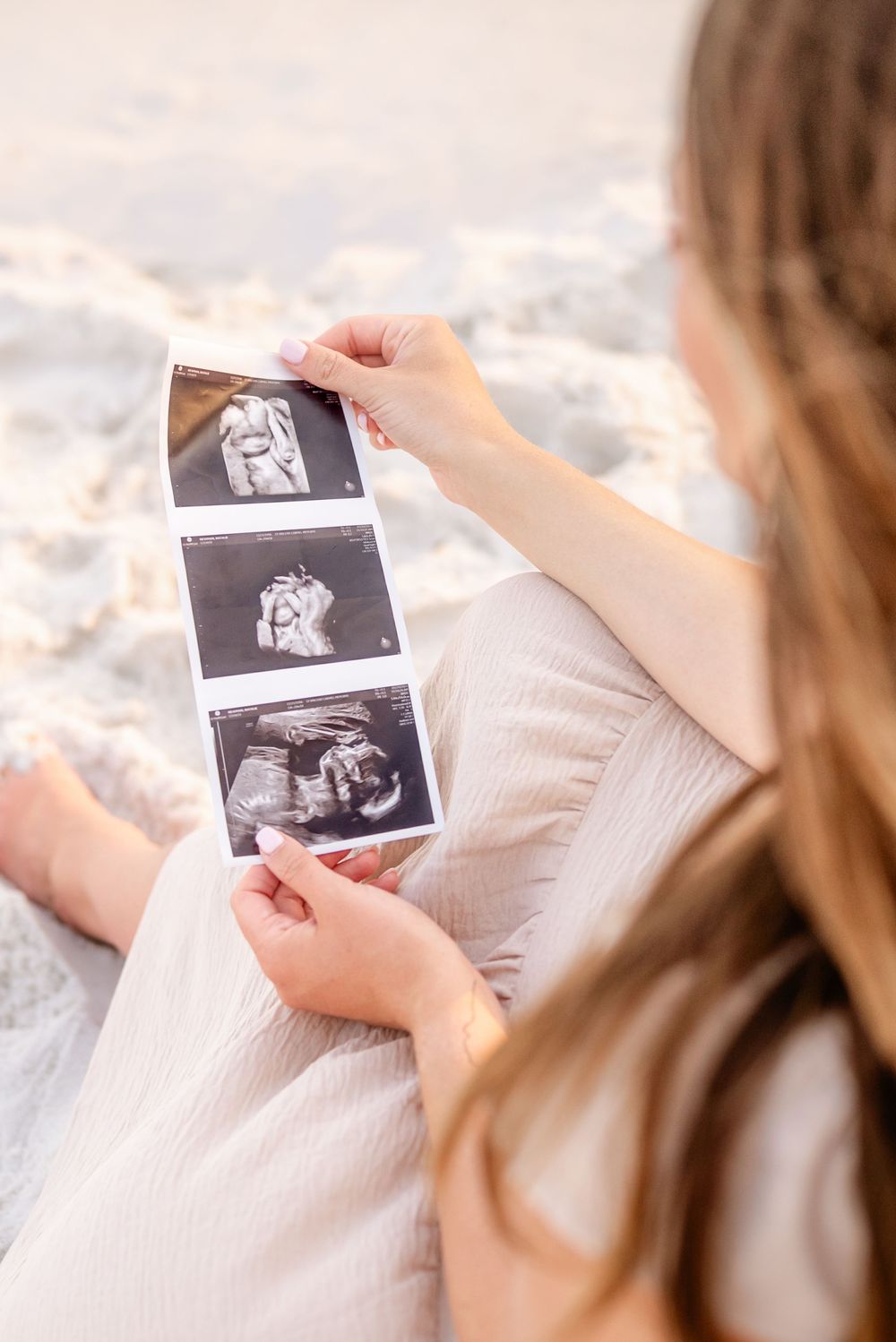 Pregnant woman sitting on sandy beach holding ultrasound photos, wearing a light pink dress.