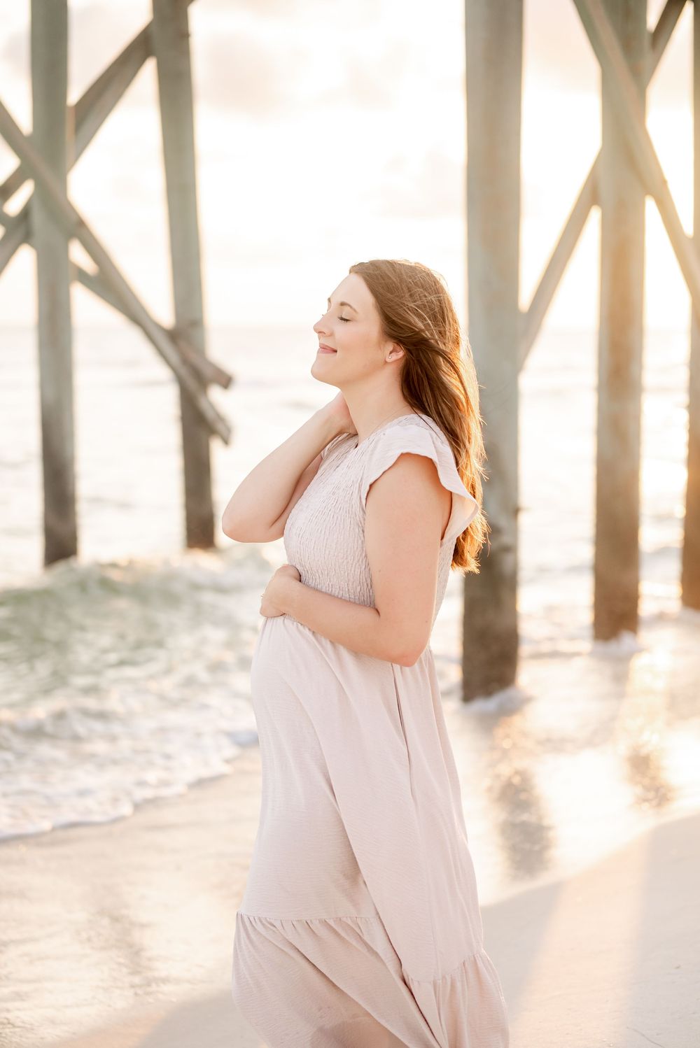 Pregnant woman in blush maxi dress smiling at beach pier during golden hour sunset.