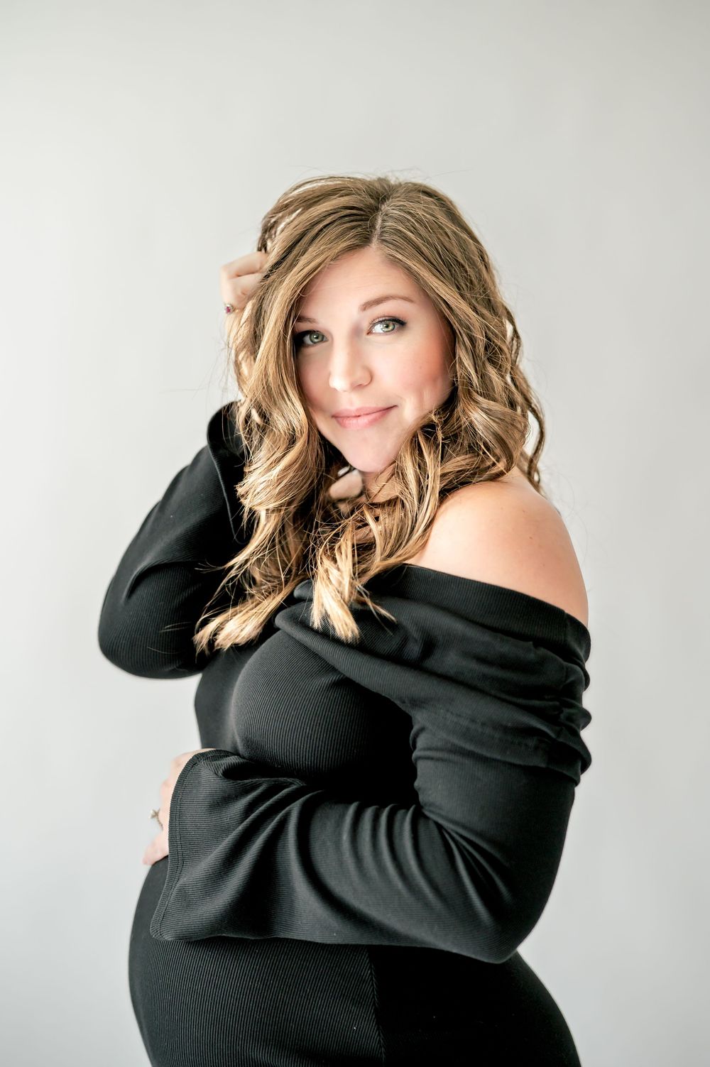 Pregnant woman in black off-shoulder dress poses gracefully, cradling baby bump with wavy blonde hair in studio.