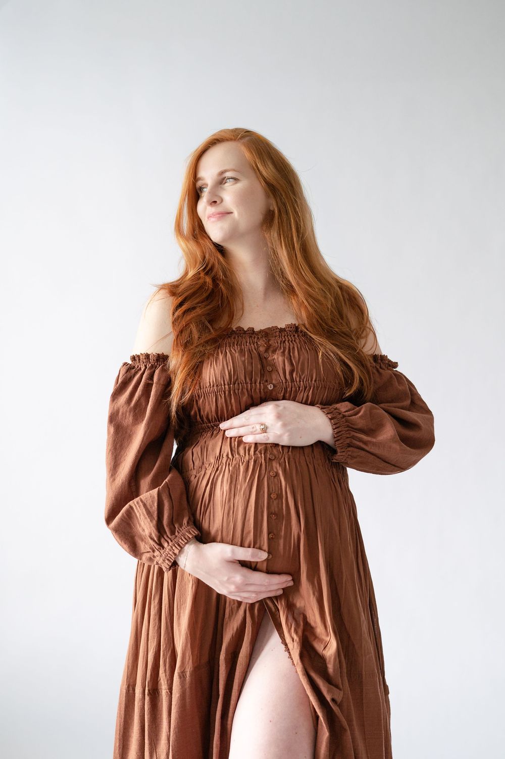 Pregnant woman in brown off-shoulder dress cradling baby bump, long red hair, studio white background.