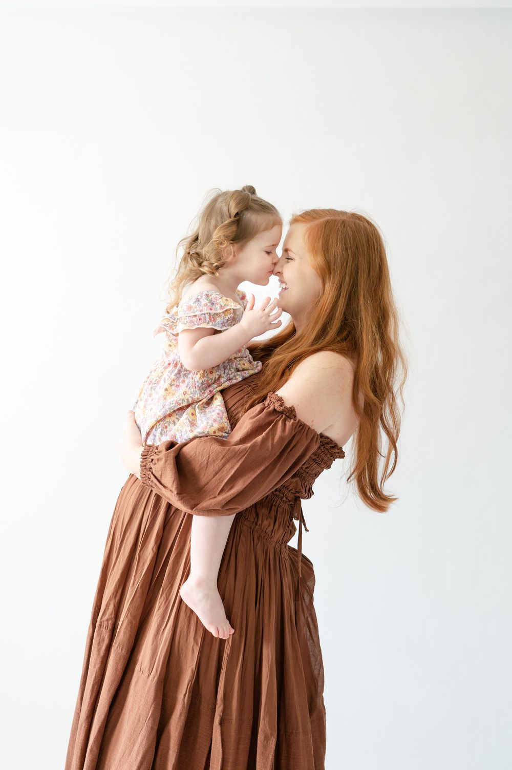 A redheaded mother in a brown dress holds her smiling toddler daughter close, touching noses in a tender moment.