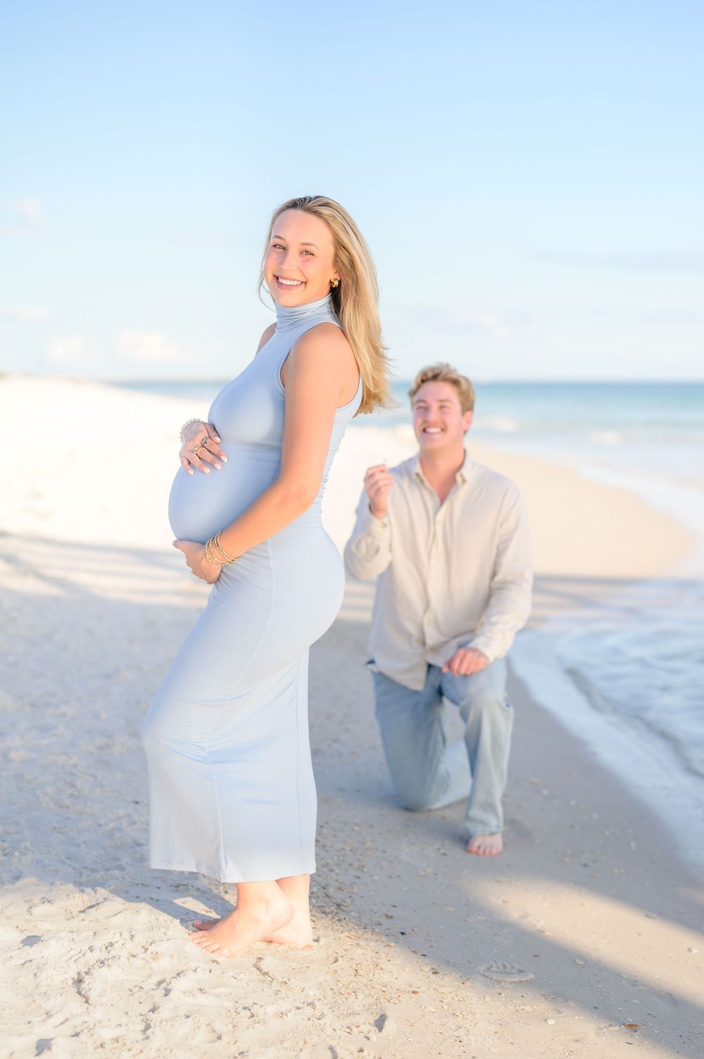 Pregnant woman in white dress holds baby bump on beach while man kneels beside her smiling during maternity photoshoot.
