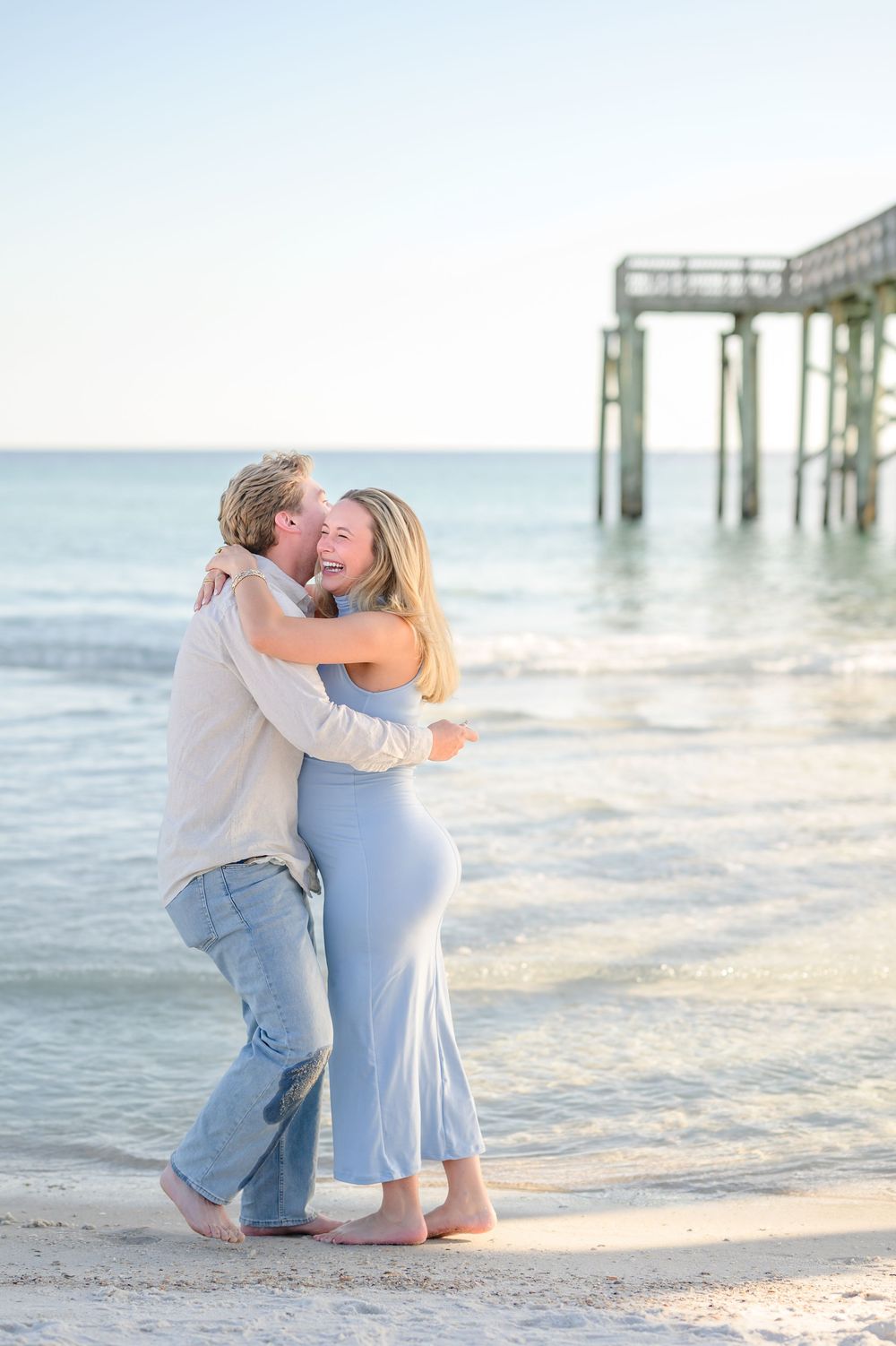 A couple embraces and laughs on a sandy beach near a pier, woman in blue dress and man in jeans.