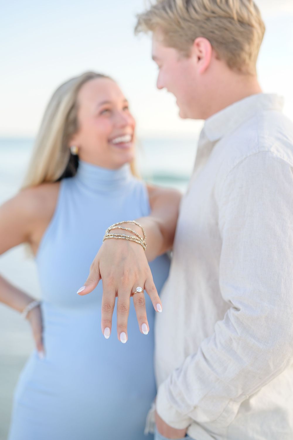 Couple at beach engagement reveal, woman showing diamond ring and bracelets, both smiling, wearing light blue and white.
