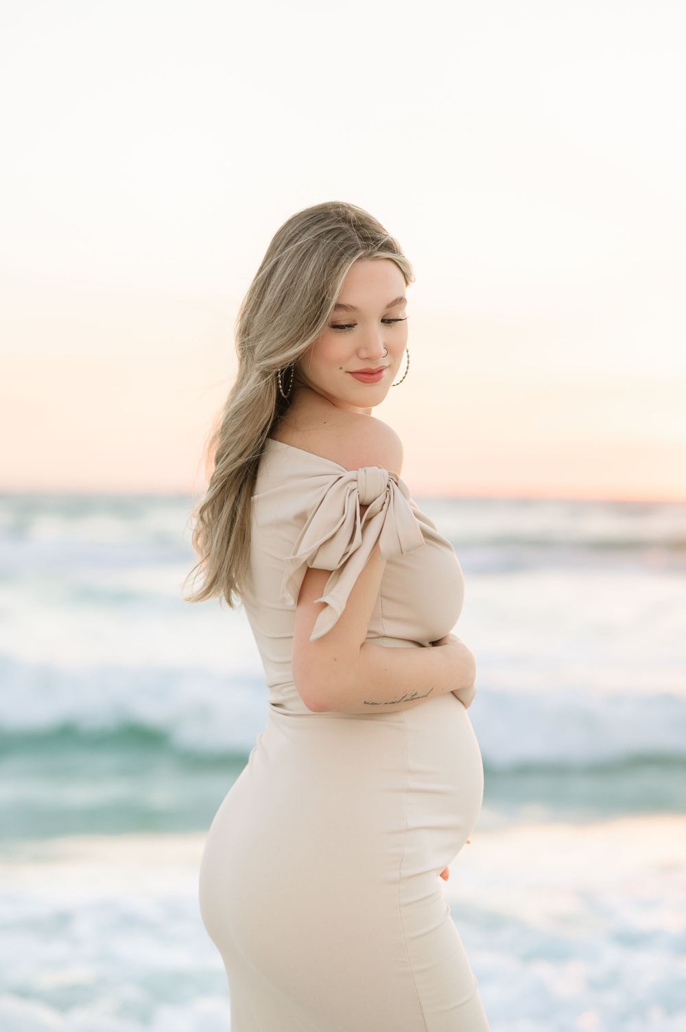 Pregnant woman in beige off-shoulder dress cradling her bump on a beach at sunset.